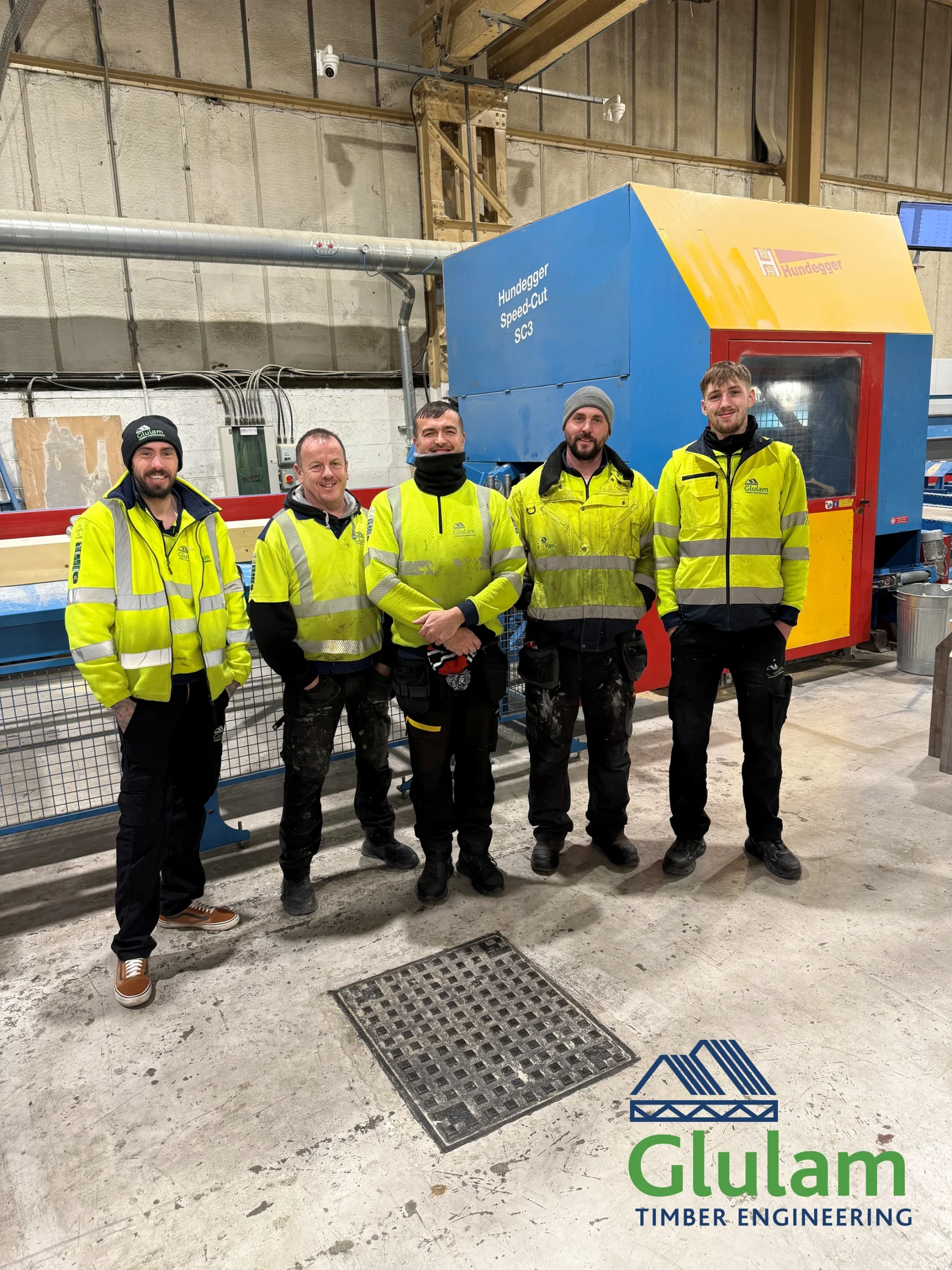 5 Glulam workers standing in front of a machine in their hi-vis smiling to the camera.